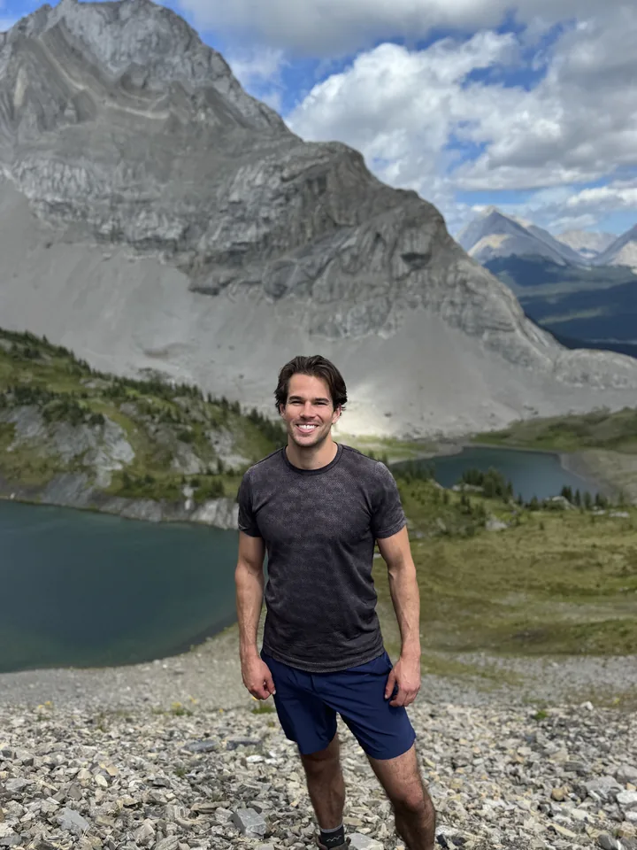 Eric smiling during a mountain hike, with a lake and alpine peaks behind him