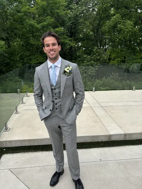 Eric standing with his hands in his pockets in a grey suit at an outdoor wedding